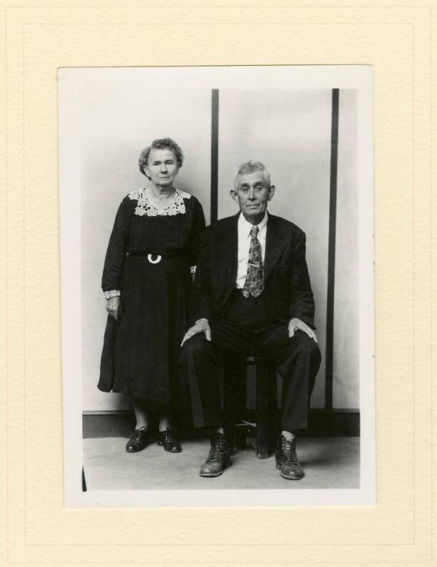 ELDERLY WELL-DRESSED COUPLE, WIFE STANDING, HUSBAND SEATED, STRIPED BACKGROUND