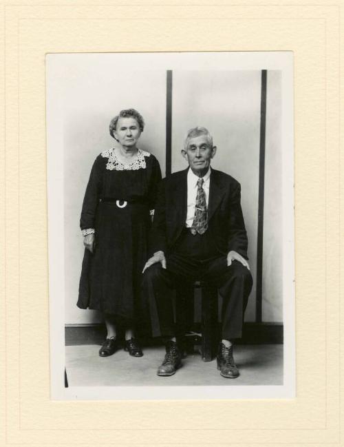 ELDERLY WELL-DRESSED COUPLE, WIFE STANDING, HUSBAND SEATED, STRIPED BACKGROUND