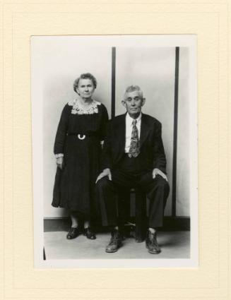ELDERLY WELL-DRESSED COUPLE, WIFE STANDING, HUSBAND SEATED, STRIPED BACKGROUND